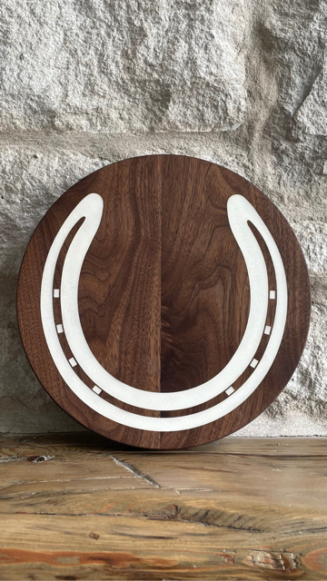 Single walnut charcuterie board with white epoxy horseshoe inlay displayed upright on a rustic wood surface in front of a textured stone wall — custom polo award by The Golden Pony.